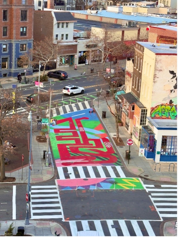 Colorful painted crosswalk and street mural at an urban intersection, surrounded by multi‑story buildings and parked cars.